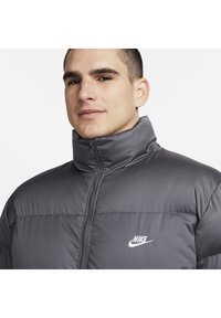 Szürke puffermellény magas gallérral, cipzárral és fehér Nike logóval a bal mellen. Simaszövet, steppelt kialakítással.