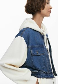 Denim jacket with a cropped design, blue fabric, front pocket, and white hoodie underneath. Smooth texture and contrasting colors.
