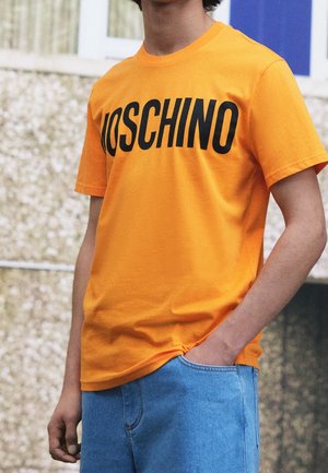 Jasnopomarańczowa koszulka z bawełny, z czarnym napisem "MOSCHINO" na piersi. Krótkie rękawy, luźny krój. Noszona z niebieskimi dżinsowymi szortami.