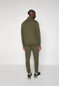 Ensemble de survêtement vert olive composé d'une veste zippée et d'un pantalon de jogging assorti, avec des poignets côtelés et une finition en tissu doux. Baskets noires incluses.