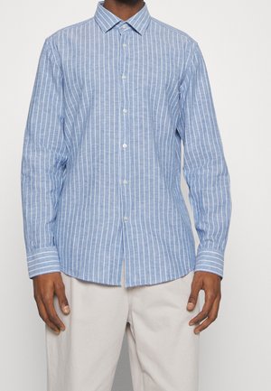 Mężczyzna w jasno niebieskiej koszuli z długim rękawem zapinanej na guziki z białymi pionowymi paskami i beżowych spodniach na jednolitym tle.