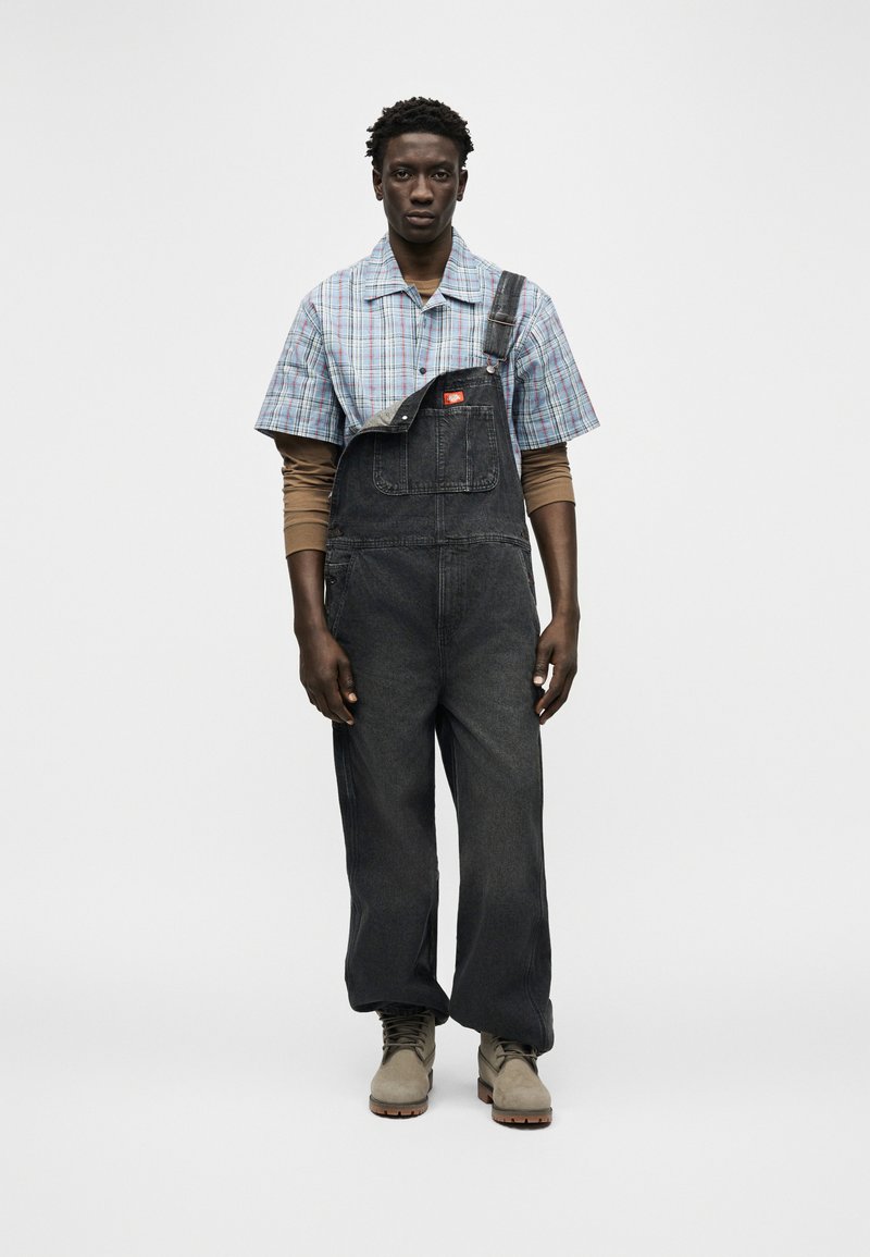 Giovane uomo in piedi che indossa una salopette di denim nero con una bretella slacciata, una camicia a maniche corte a quadri blu sopra una maglia marrone a maniche lunghe e stivali beige.