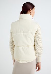 Gilet matelassé de couleur crème avec un col haut et des manches texturées en beige. Tissu doux avec un design matelassé et un ourlet élastique.