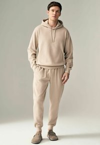 Hanorac bej cu glugă, dotat cu buzunar frontal și șnur de ajustare, asortat cu pantaloni de trening tapered. Textură moale cu manșete elastice.
