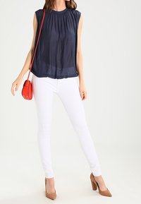 Kvinna klädd i ett ärmlöst marinblått linne, vita slim fit-jeans, bruna högklackade skor och bär en liten röd axelväska.