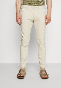 Beige chinos med slim fit, der har sidelommer og oprullede manchetter, parret med brune ruskinds sandaler. Stoffet ser let og glat ud.