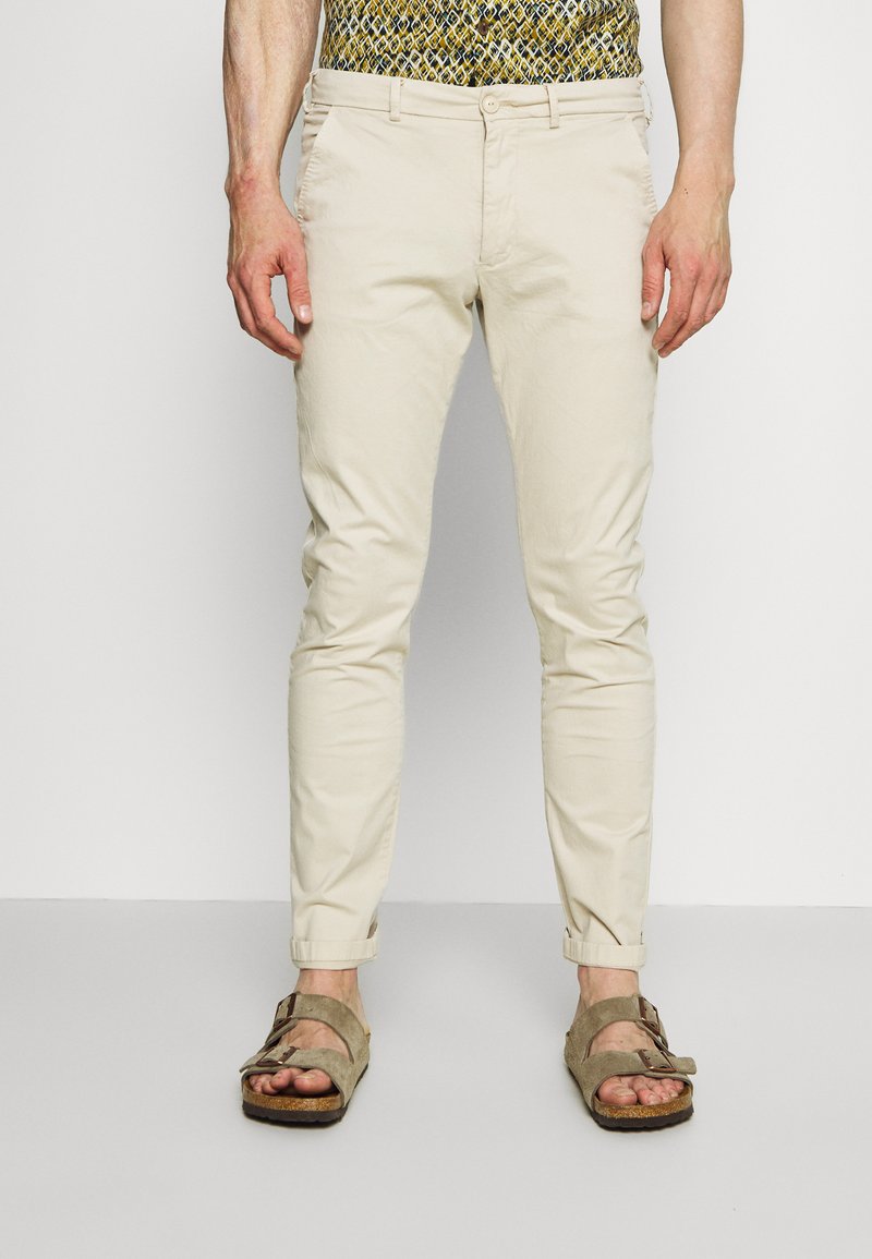 Beige chinos med slim fit, der har sidelommer og oprullede manchetter, parret med brune ruskinds sandaler. Stoffet ser let og glat ud.