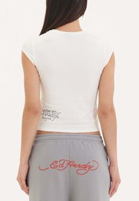 Bílý zkrácený tričko s krátkým rukávem, na zadní straně s černým logem Ed Hardy, kombinované s šedými tepláky s červeným logem.