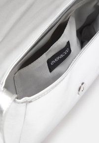 Hopeapintainen käsilaukku, jossa on sileä nahkapinta, napituskiinnitys ja harmaa kankainen sisustus, jossa on sisätasku, jonka etiketissä lukee "even&odd."