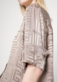 Blusa marrone a fantasia con una texture seta, caratterizzata da un design geometrico e maniche arrotolate, con dettaglio di bottoni sui polsini.