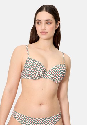 Mujer con un bikini de estampado a rayas zigzag en verde, blanco y naranja, posando frente a un fondo liso de color gris claro.