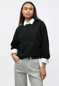 Γυναίκα με μακριά σγουρά μαλλιά, φορώντας ένα μαύρο πλεκτό πουλόβερ με γράμματα "HUGO", λευκό πουκαμισάκι και ανοιχτό γκρι τζιν, ποζάροντας με το χέρι στην τσέπη.