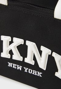 Sac en toile noir avec des accents en cuir blanc et une inscription en relief "KNY NEW YORK." Présente une ouverture zippée et des poignées robustes.