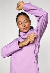Fioletowa wodoodporna kurtka z wysokim kołnierzem, zapinanymi na zamek kieszeniami bocznymi i regulowanymi mankietami. Gładka tekstura i sportowy design.