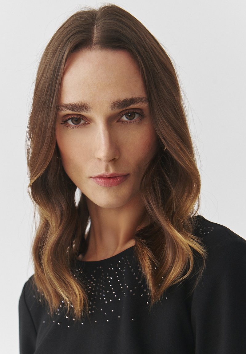 Czarna odzież z okrągłym dekoltem, z błyszczącymi ozdobami z przodu. Długie, falujące włosy opadają na ramiona.