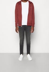 Man med en röd zip-up hoodie, vit t-shirt, blekta svarta jeans och vita sneakers som står mot en enkel vit bakgrund.