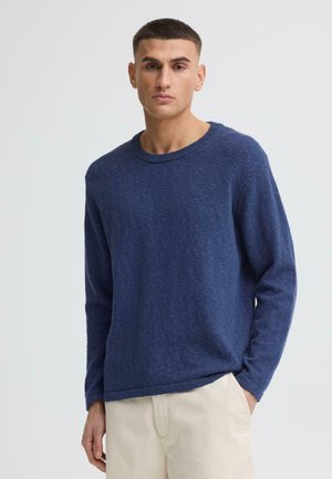 Junger Mann mit kurzen Haaren, der einen blauen Pullover mit langen Ärmeln und hellbeige Hose vor einem schlichten hellen Hintergrund trägt.