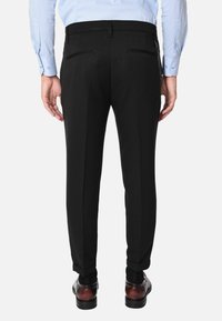 Pantaloni neri con una vestibilità su misura, dotati di due tasche posteriori, cerniere discrete e una texture liscia; indossati con scarpe eleganti marroni.