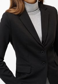 Femme portant un blazer noir ajusté à deux boutons sur un pull à col roulé gris clair, raccourci au niveau des épaules et de la taille.
