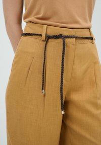 Beige broeken met een textuurstof, voorzien van een gevlochten bruin leren riem met metalen uiteinden, gestyled met een hoge taille en plissé aan de voorkant.