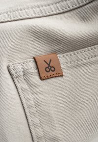 Beige stofstaal met een bruine leren label met schaarlogo, gestikt langs de rand, dat een gestructureerd oppervlak toont.