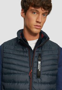 Marineblauer Puffer-Weste mit gestepptem Design, Reißverschluss und kontrastierendem Innenkragen. Mit roten Reißverschluss-Akzenten und einem "Elevated Casual"-Label.