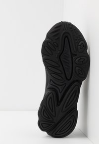 Černá podrážka tenisky s texturovaným vzorem, obsahující drážky a vyvýšené akcenty pro lepší grip, pohled zespodu na bílém pozadí.