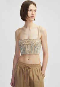 Beaded crop top met dunne spaghettibandjes, met een beige basis en ingewikkelde witte en zilveren kralenwerk in abstracte patronen.