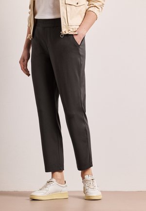 Osoba w czarnych zwężanych spodniach z elastycznym pasem, beżową kurtce, białych trampkach, stojąca na tle jednolitego jasnego tła.