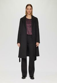 Zwarte lange jas met een reverskraag, twee knopen aan de voorkant en een strikdetail aan de zijkant, gedragen over een gemusterde donkere top en zwarte broek. Glanzende zwarte schoenen.