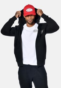 Uomo che regola la felpa nera su una t-shirt bianca Von Dutch e un cappellino rosso Von Dutch, su uno sfondo chiaro e semplice.