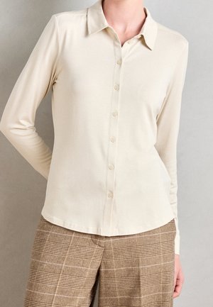 Persoon die een crèmekleurig langmouwen overhemd met knopen draagt, gecombineerd met een bruine geruite broek, staand tegen een effen lichtgrijze achtergrond.