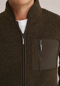 Chaqueta de forro polar marrón con un alto cuello, que cuenta con un bolsillo con cremallera en el pecho, tejido texturizado y acentos suaves en contraste.