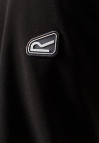 Tissu noir avec un petit patch triangulaire noir et blanc présentant une lettre « R » stylisée au centre.