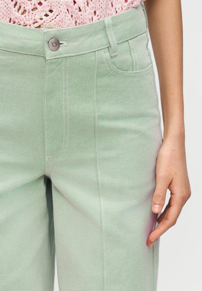 Κοντινό πλάνο ενός ατόμου που φοράει ανοιχτό πράσινο τζιν παντελόνι και ροζ πλεκτό πάνω, εμφανίζοντας κουμπί, τσέπες και μέρος του δεξιού του χεριού.