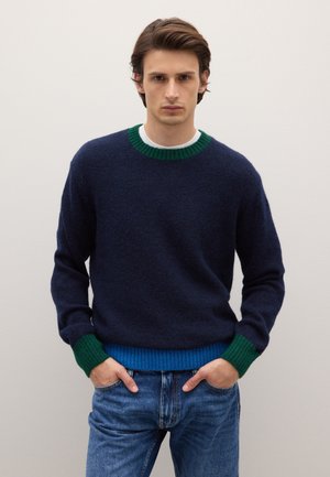 Marineblauer Strickpullover mit grünen und blauen, gerippten Bündchen an Ärmeln und Saum. Rundhalsausschnitt. Das Modell trägt ihn mit blauen Denim-Jeans. Glatter, strukturierter Stoff.