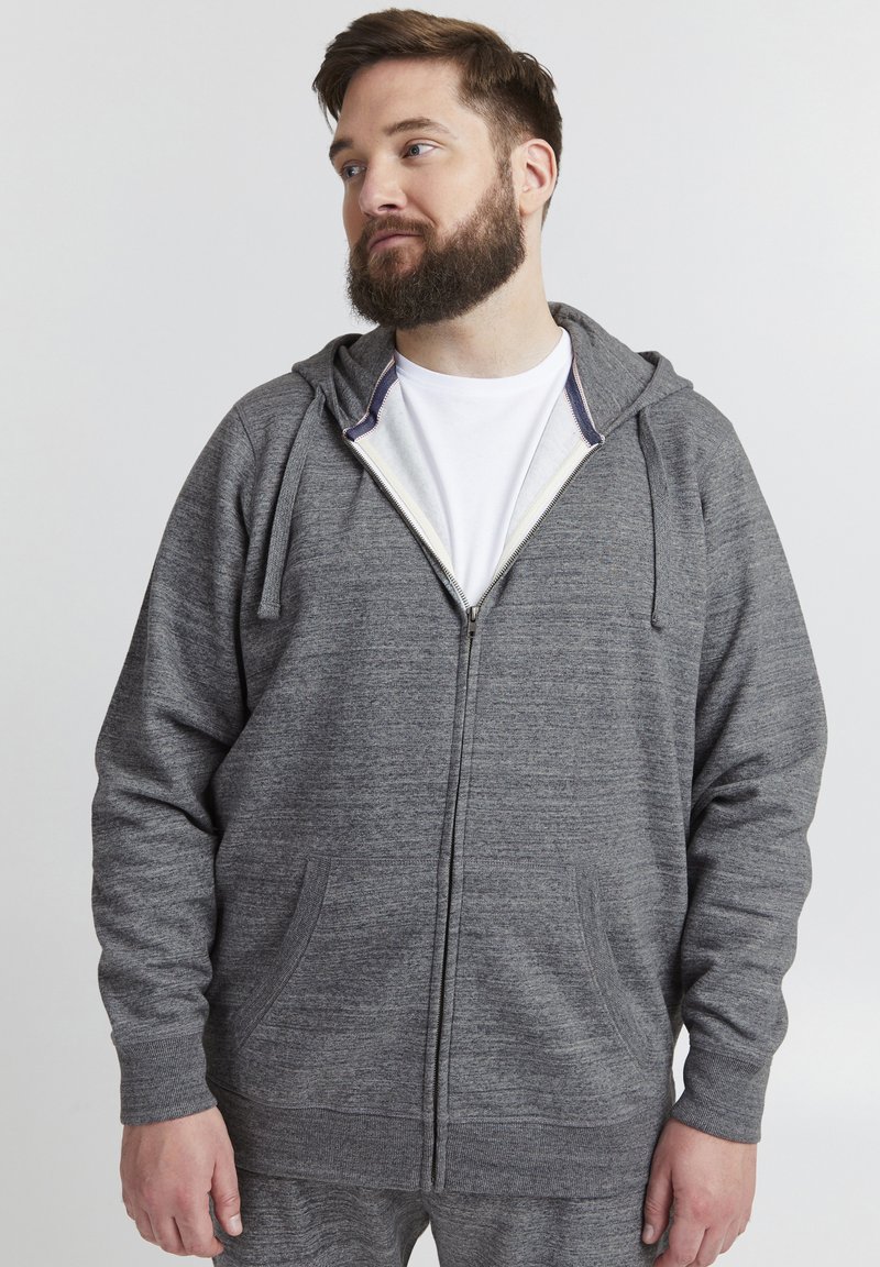 Siva zip-up dukserica s teksturiranim materijalom, dva prednja džepa i podrživač za podešavanje kapuljače. Sadrži bijelu podstavu blizu zatvarača.