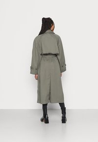 Zielona oliwkowa trencz z krótkim krojem, zwiniętymi rękawami i paskiem do związania, wykonany z gładkiego materiału. Na nogach są czarne botki na obcasie.