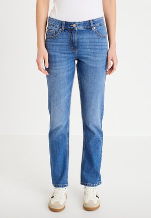 Donna che indossa jeans blu a gamba dritta di lavaggio medio con tasche frontali, vita con bottoni, sneaker bianche e una maglietta bianca su uno sfondo semplice.