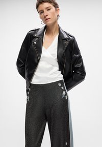 Veste de motard en faux cuir noir avec des fermetures éclair argentées, associée à un haut wrap en satin blanc et un pantalon noir à effet métallique avec broderies florales.
