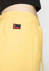 Gelbe Sweatpants aus weichem Stoff, mit einem schwarzen quadratischen Patch und einem roten 'L'-Logo sowie seitlichen Taschen zur Aufbewahrung.