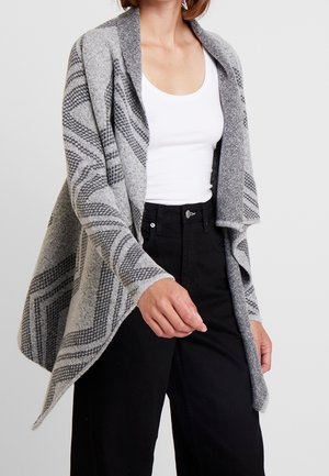 Frau trägt eine graue, gemusterte Strickjacke mit offenem Vorderteil über einem weißen Tanktop und hoch taillierten schwarzen Hosen, steht vor einem schlichten Hintergrund.