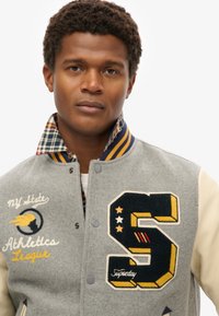 Grijze wolmix varsityjack met crèmekleurige mouwen, geborduurd "S" embleem, decoratieve stiksels, geruite voering en drukknoopsluiting.