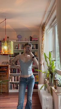 Camisola ajustada azul-claro com rendas, combinada com jeans azuis flare. O fundo apresenta prateleiras de livros e uma lâmpada pendurada.