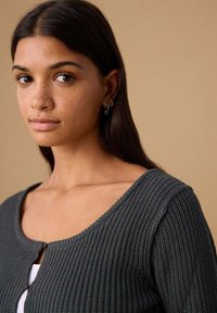 Dunkelgrauer Rippsweater mit V-Ausschnitt, verfügt über einen Knopfverschluss und eine strukturierte Stoffoberfläche. Das Modell trägt kleine Creolen vor neutralem Hintergrund.