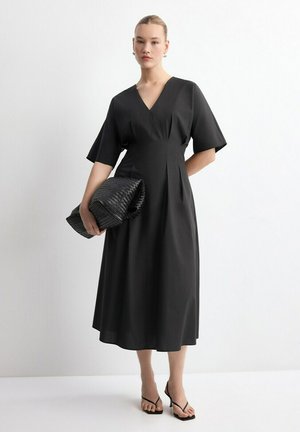 Mujer con un vestido midi negro de escote en V y mangas anchas, sosteniendo un gran clutch tejido negro, posando sobre un fondo blanco.