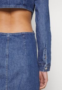 Denimová dvoudílná souprava obsahující cropped bundu s dlouhými rukávy a úzkou sukni, obojí v středním odstínu modré s viditelným prošíváním a detaily kovových knoflíků.