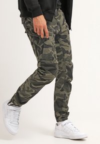 Spodnie typu cargo w ciemnozielonym camo, lekka tkanina, zwężany fason, liczne kieszenie, zestawione z białymi trampkami.