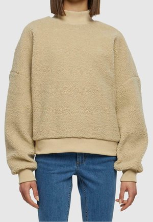 Beige fleecetrui met hoge kraag, verlaagde schouders en geribde zoom. Zacht, текстuurlijk materiaal met een relaxte pasvorm, gecombineerd met blauwe jeans.