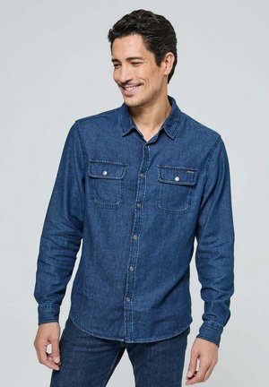 Chemise en denim bleu foncé, dotée de manches longues, d'un devant à boutons, de deux poches sur la poitrine et de coutures contrastantes. Texture lisse.